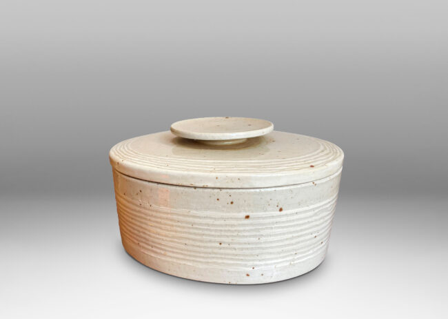 Gallery BAC oval form, with carved ridges encircling the box and bordering the edges of the lid; hand-crafted in a speckled pale gray stone ware