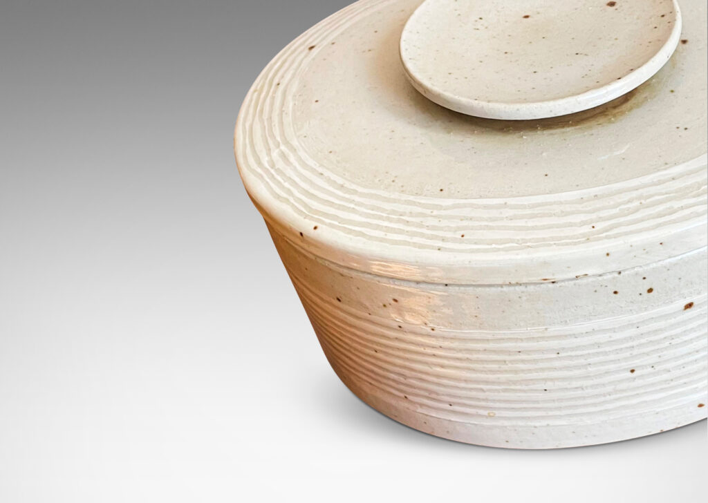 Gallery BAC oval form, with carved ridges encircling the box and bordering the edges of the lid; hand-crafted in a speckled pale gray stone ware