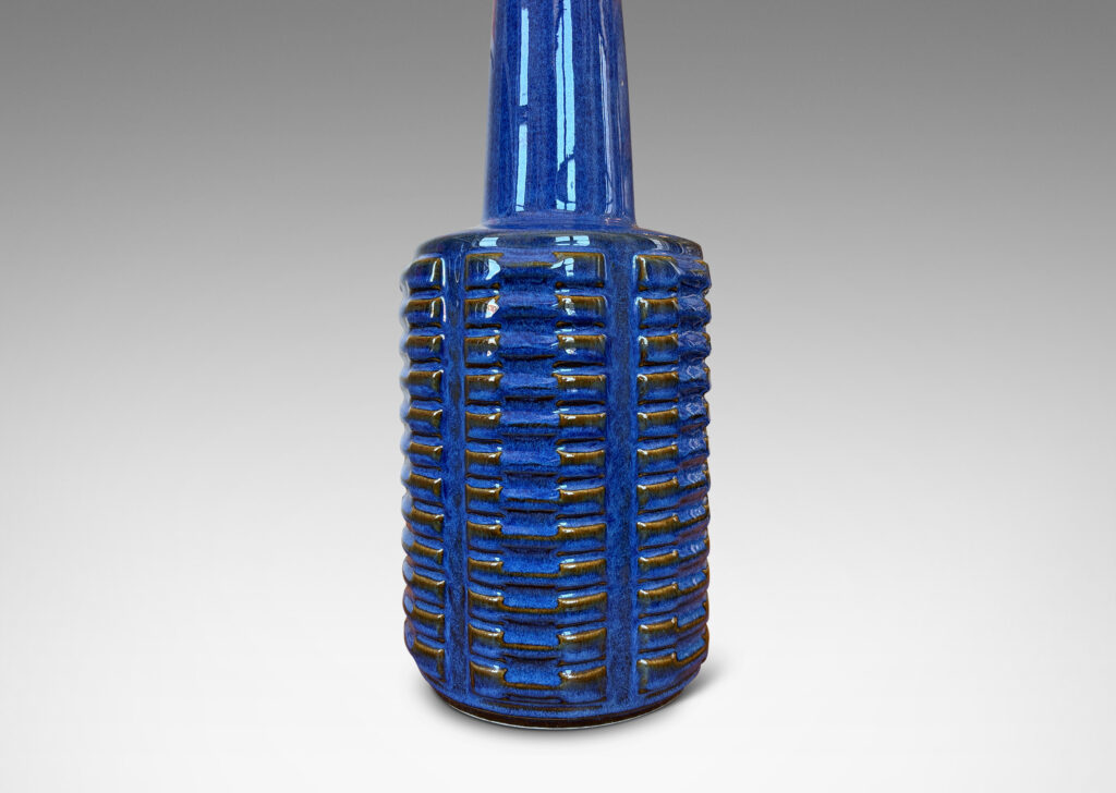 Gallery BAC cylindrical form with stacks of impressed motifs in relief and a tapered neck, glazed in a saturated blue-brown