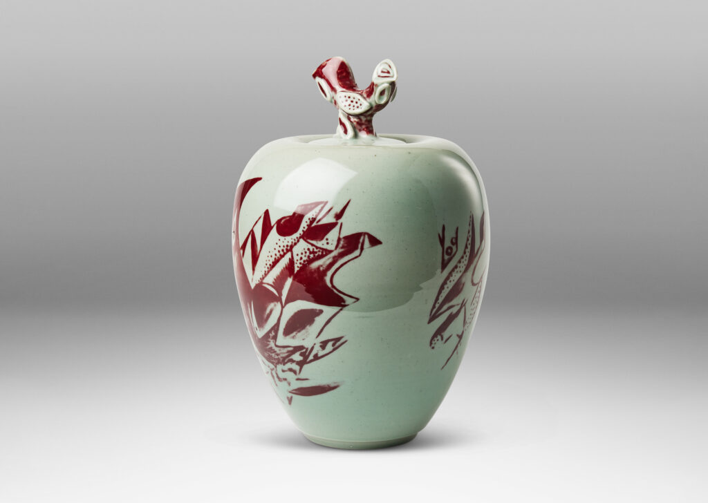 Gallery BAC tapered form and lid with branch-like finial, glazed in celadon with hand-painted abstracted fish, plants, shells and patterning in oxblood