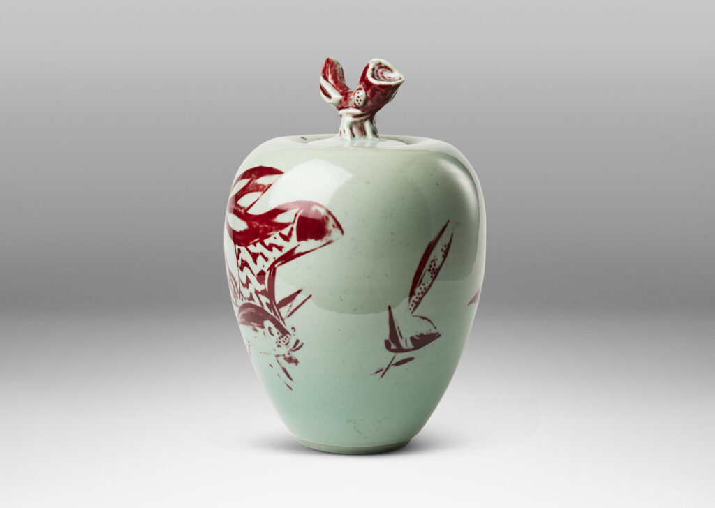 Gallery BAC tapered form and lid with branch-like finial, glazed in celadon with hand-painted abstracted fish, plants, shells and patterning in oxblood