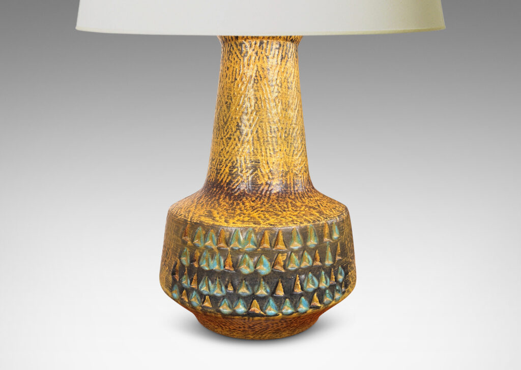 Gallery BAC floating drum form with wide conical neck, covered in a carved texture and ornamented with triangle motifs, and glazed in a mustard gold tone, with dark gray and turquoise elements