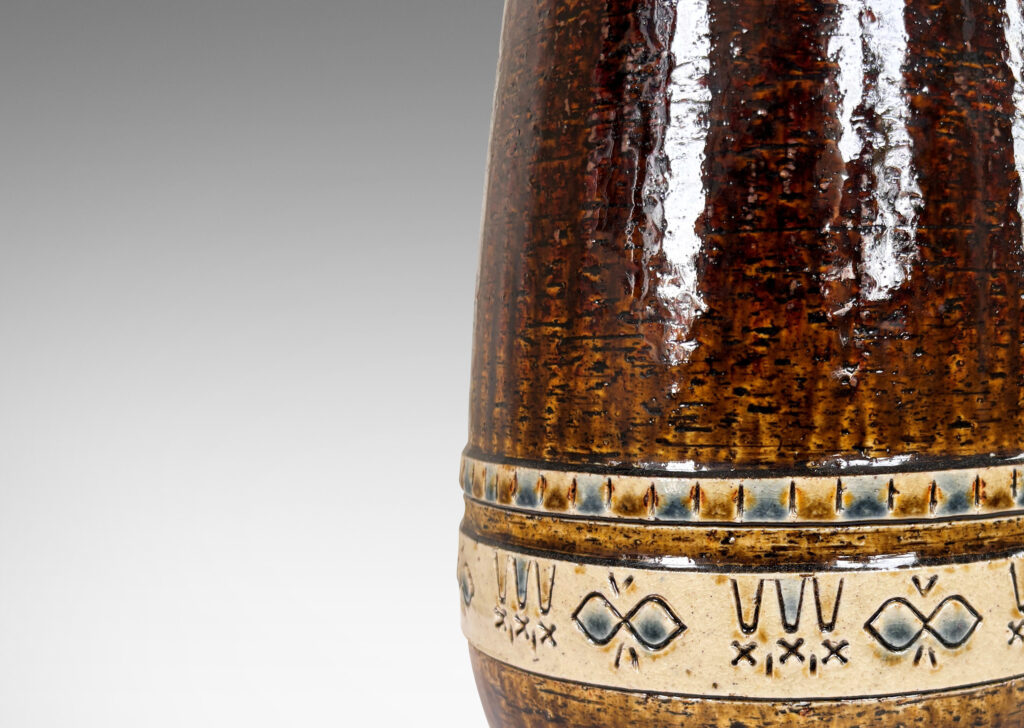 Gallery BAC having grog-textured ovoid cylindrical forms in shiny warm brown with bands of carved geometric designs in ivory, gold and blue