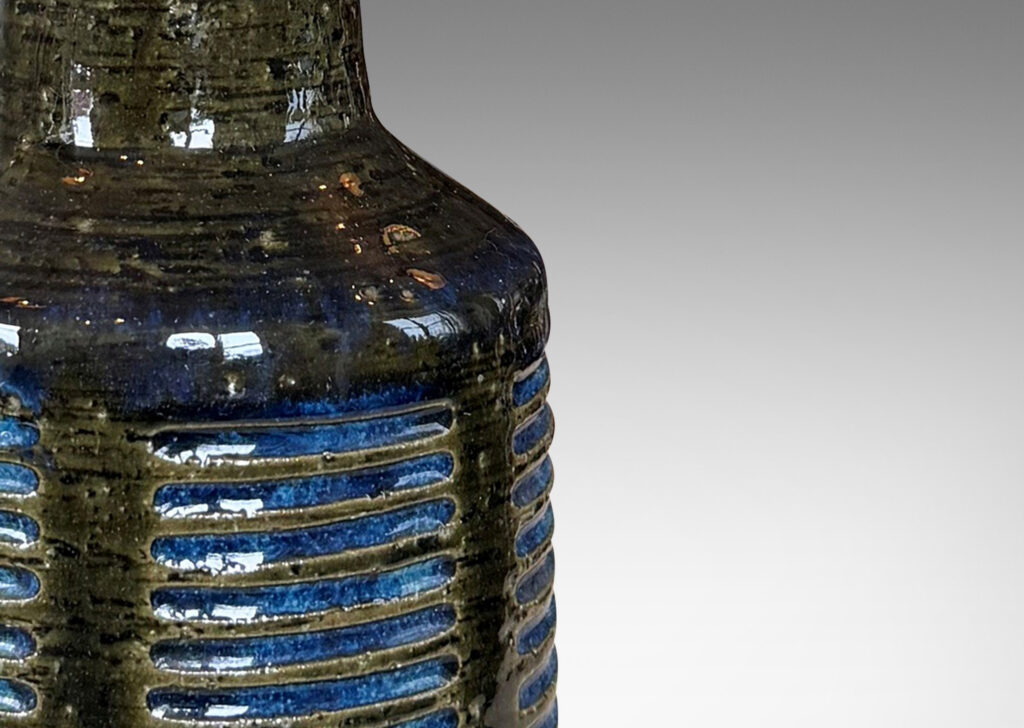 Gallery BAC drum form with vertical bands of carved lines, and tall conical neck, in a grog texture; glazed in gloss dark green with blue