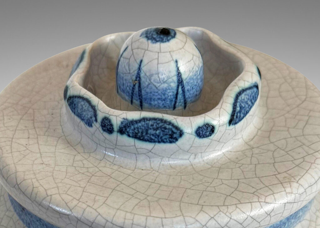 Gallery BAC undulating ovoid form with a disk lid with floral style finial, glazed in a pale gray craquel, with wildflower ornaments painted in blue